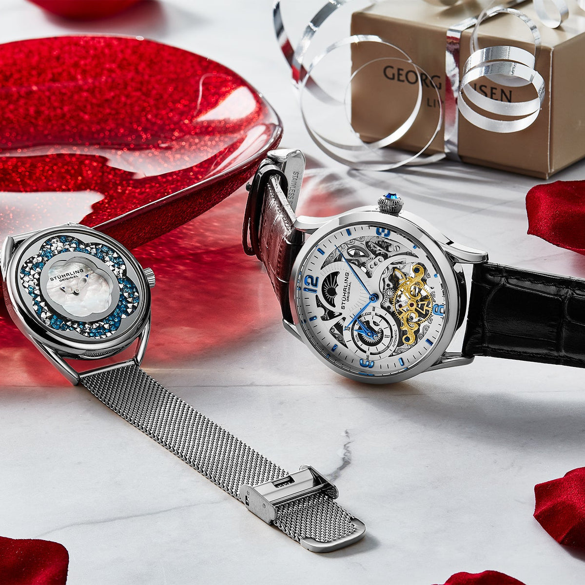 Two watches on a white surface with red petals and a box in the background